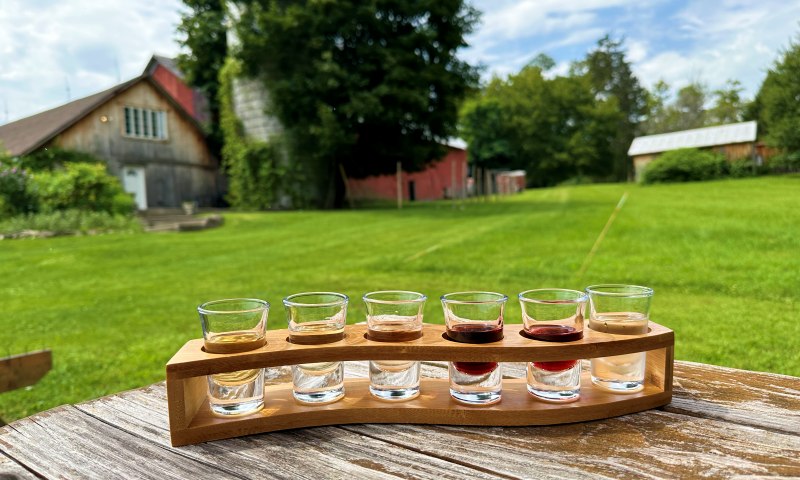 6 shot glasses of wine ina wooden stand with lawn and farmhouse in background at Baldwin Vineyards in Hudson Valley